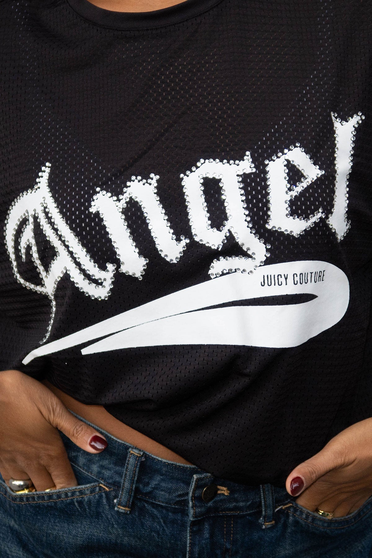 A person wears the Angel Couture Oversized Tee Dress—a black mesh style with “Angel” in bold white letters and a Juicy Couture logo—paired with blue jeans. Their hands rest on their waist, partially visible at the bottom.