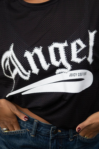 A person wears the Angel Couture Oversized Tee Dress—a black mesh style with “Angel” in bold white letters and a Juicy Couture logo—paired with blue jeans. Their hands rest on their waist, partially visible at the bottom.