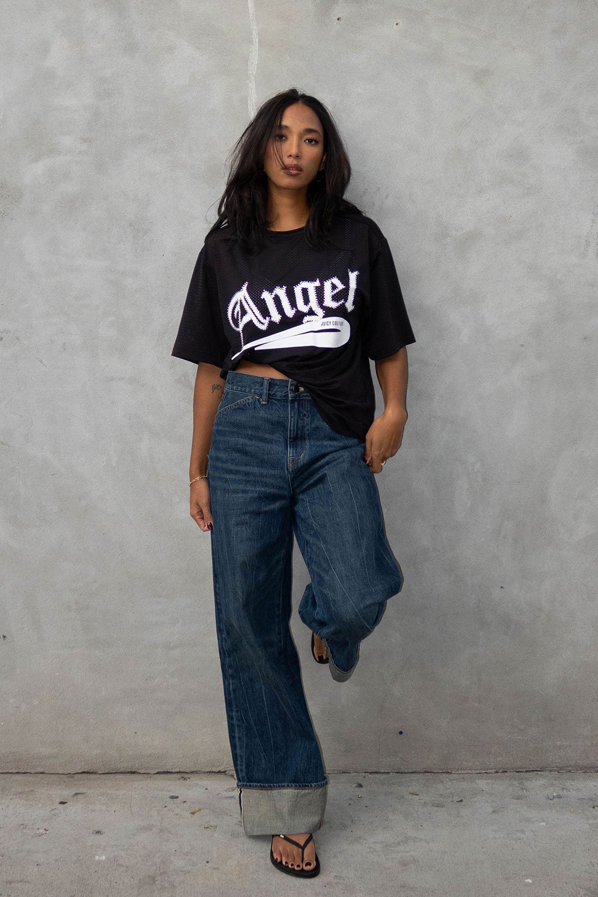 A woman with long dark hair poses confidently against a gray wall, wearing the Angel Couture Oversized Tee Dress in black with "Angel" lettering, paired with cuffed wide-leg jeans and black sandals.