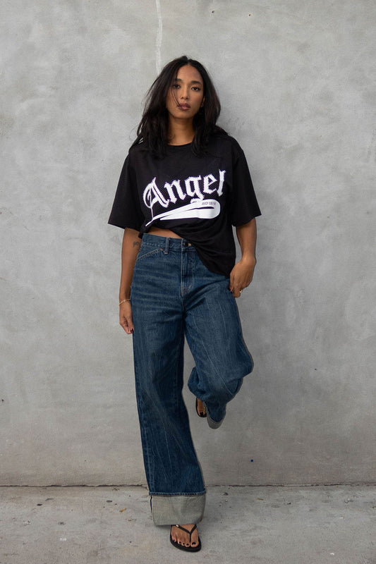 A woman with long dark hair poses confidently against a gray wall, wearing the Angel Couture Oversized Tee Dress in black with "Angel" lettering, paired with cuffed wide-leg jeans and black sandals.