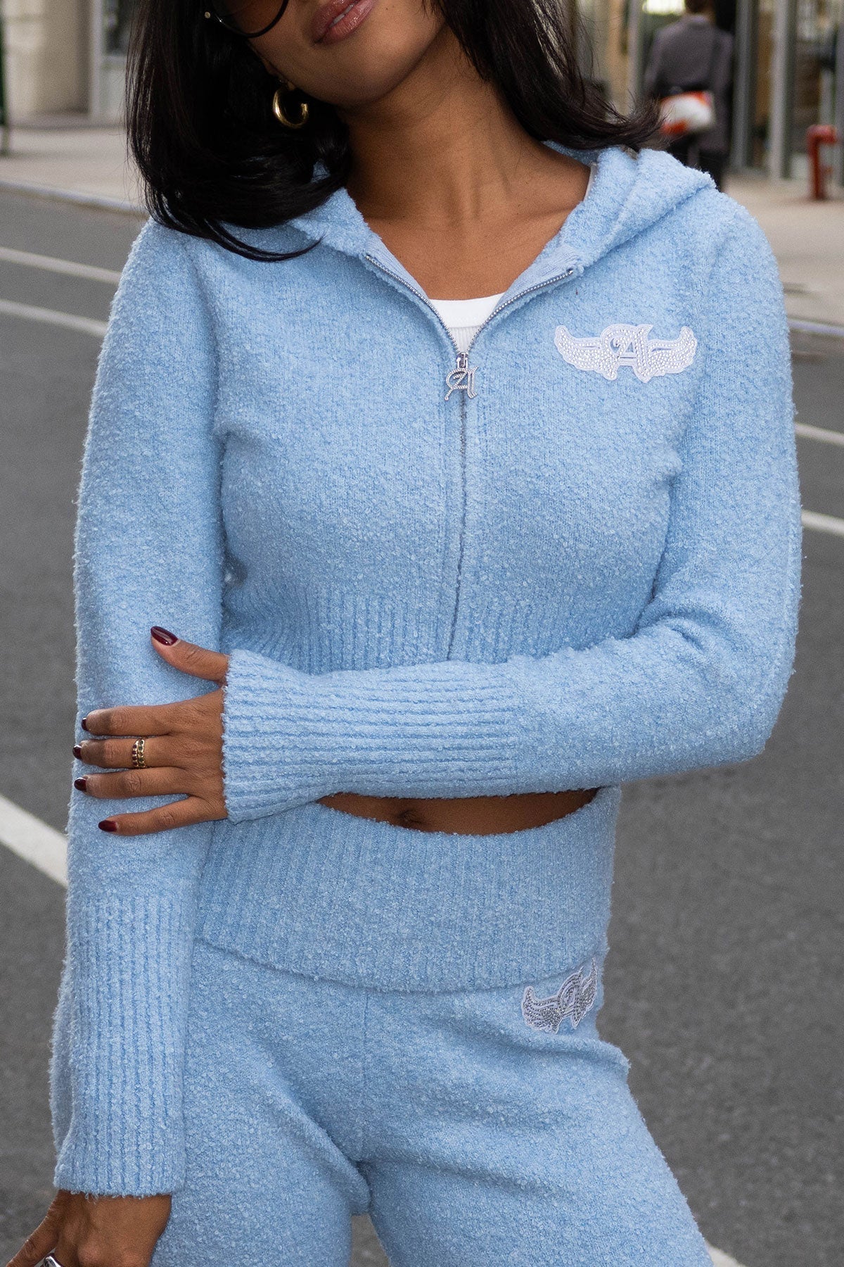Wearing the Angel Couture Knitwear Patch Hoodie from the Juicy Couture x Angel Reese collection, featuring a sparkling winged A logo, and matching pants, a person stands on a city street with arms crossed to reveal a white top underneath.