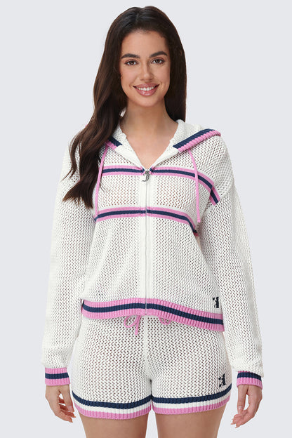 A woman stands smiling against a light gray background, wearing sporty-chic mesh shorts featuring pink and navy stripes, paired with a white knit hooded zip-up jacket.