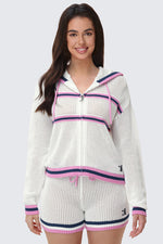 A woman stands smiling against a light gray background, wearing sporty-chic mesh shorts featuring pink and navy stripes, paired with a white knit hooded zip-up jacket.