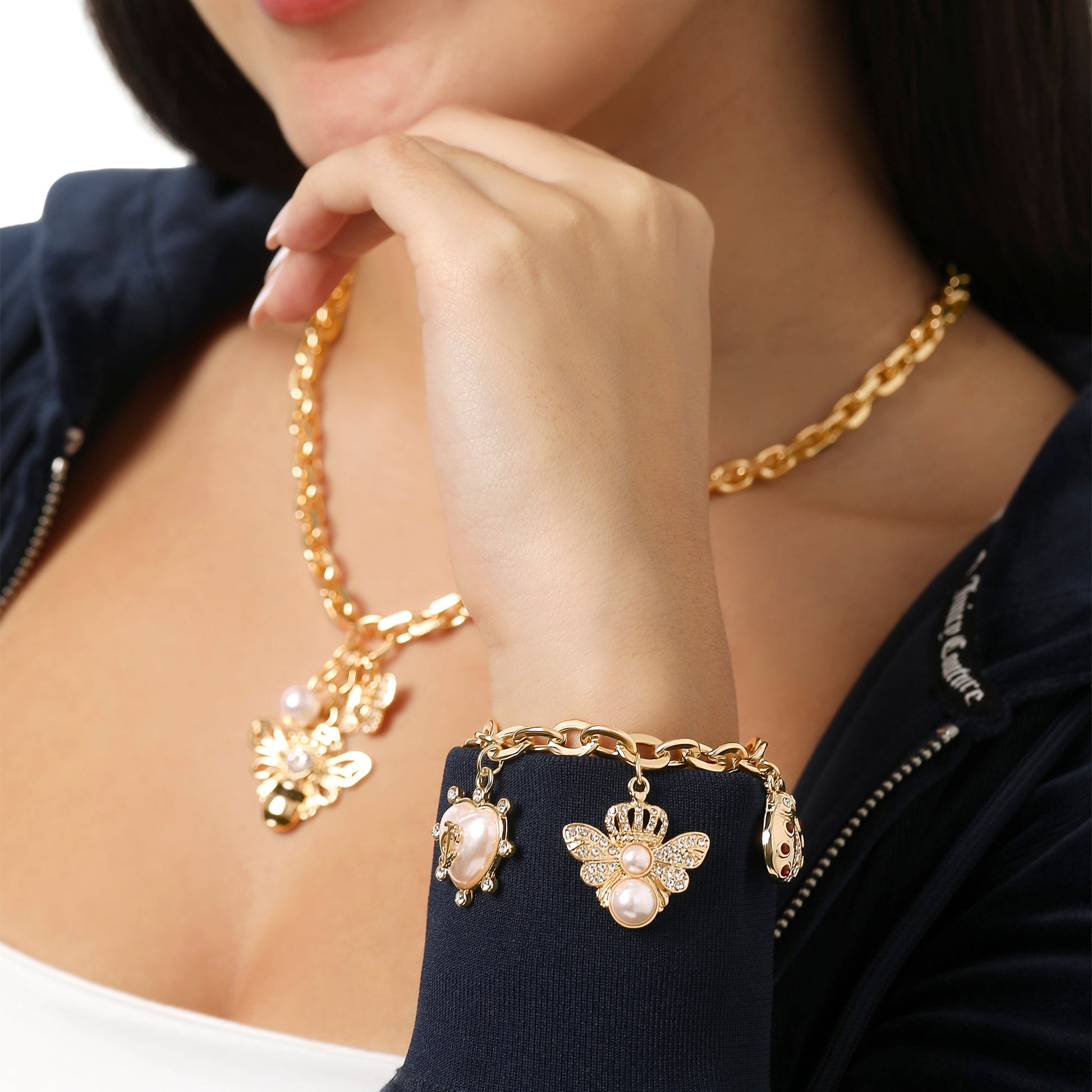 A woman in a dark jacket wears a gold chain necklace with a butterfly pendant and the Pearl & Icon Charm Bracelet, an adjustable gold-tone piece featuring pearls and butterfly charms, as she holds her hand near her chin.