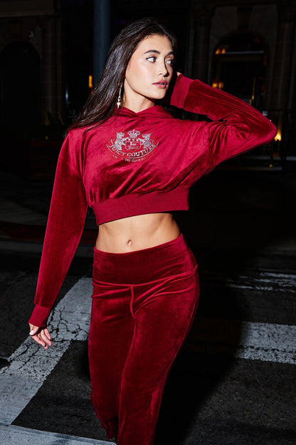 A woman wearing the Cropped Scottie Hoodie in juicy red velour with a matching skirt walks across a city street at night, glancing to the side and touching her hair.